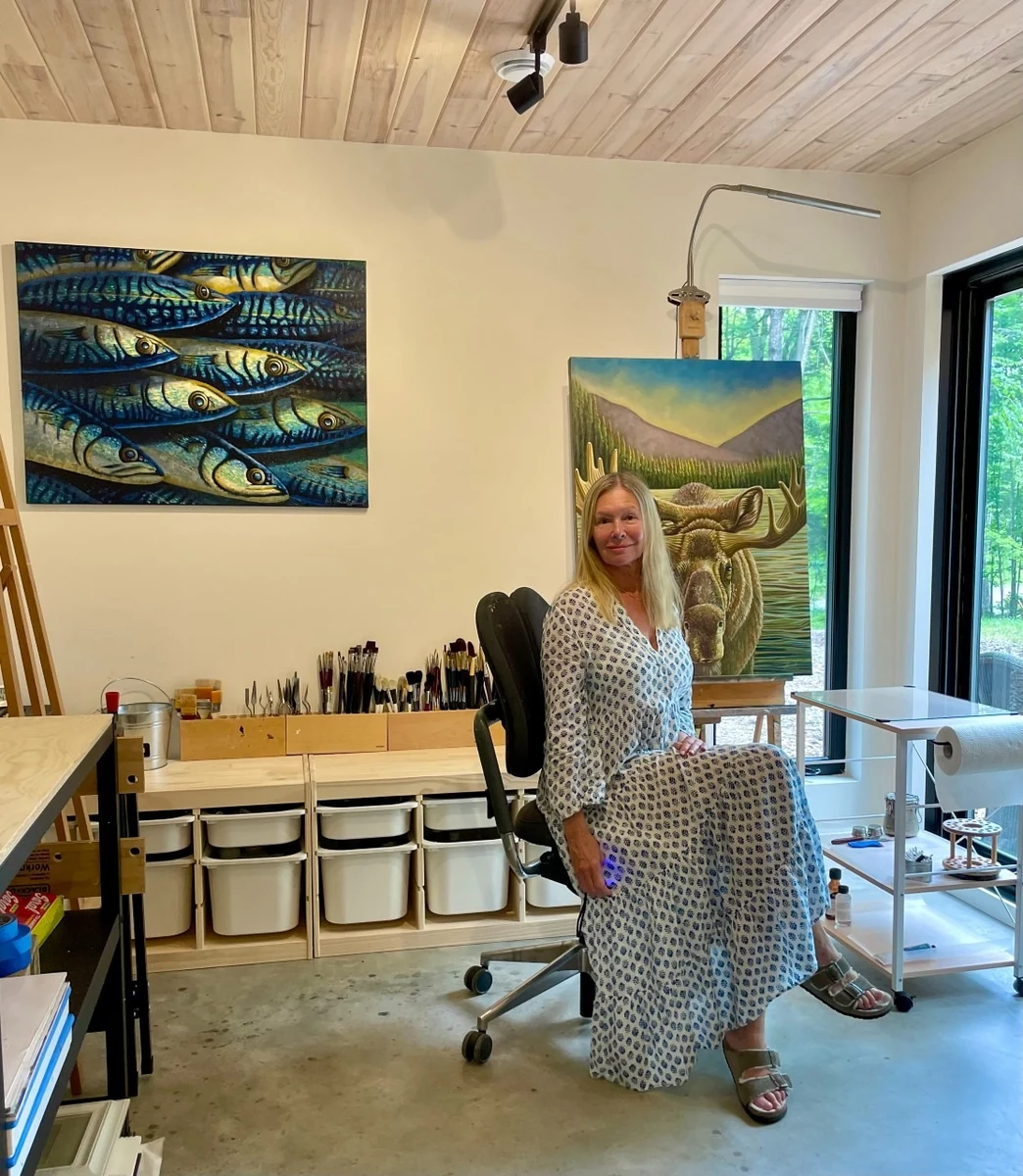 Alicia Zemanek seated in her studio, with a painting of a school of fish on the wall behind her and a moose painting on the easel