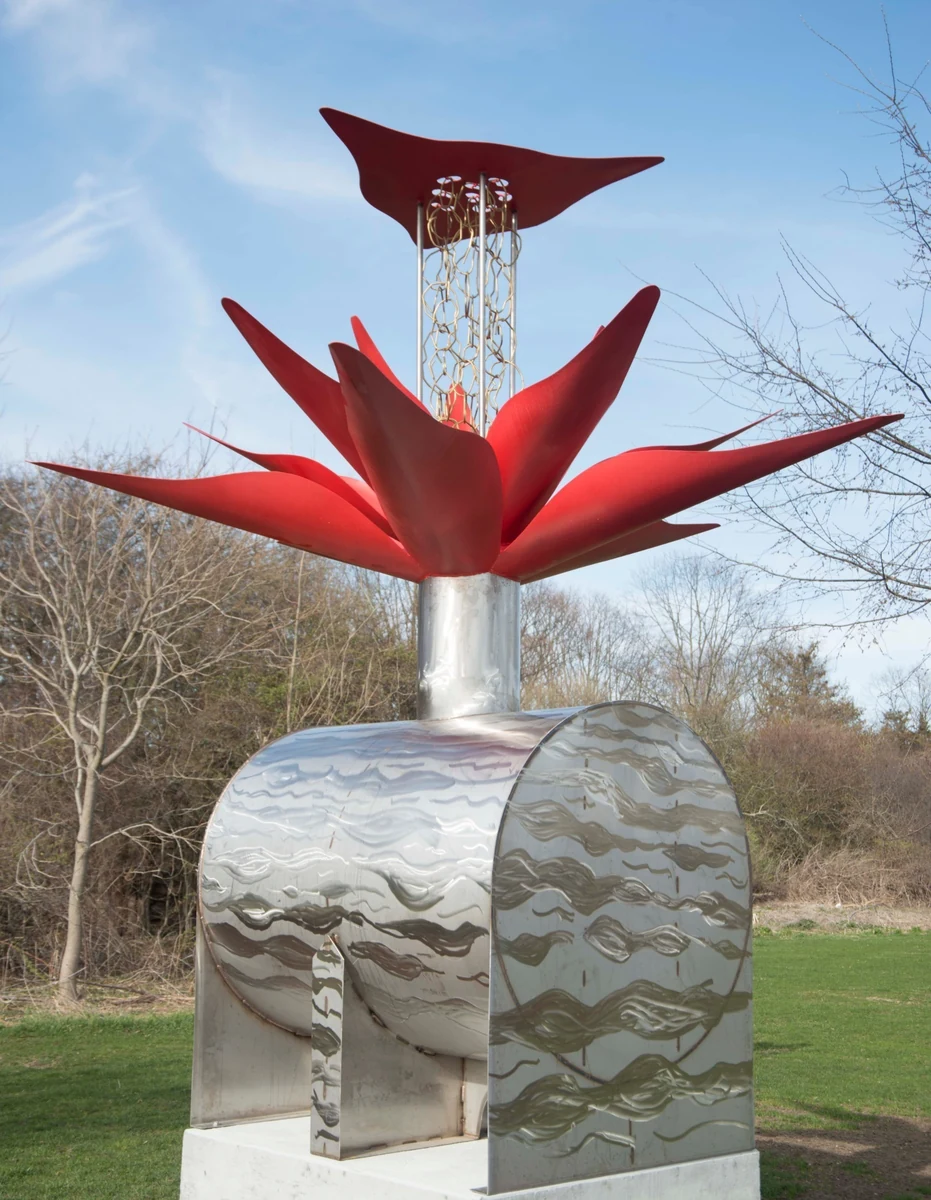 RainKeep rain-harvesting sculpture: red powder-coated petal canopy with a bronze rain chain above a polished stainless steel collection vessel, standing on a lawn against a blue sk