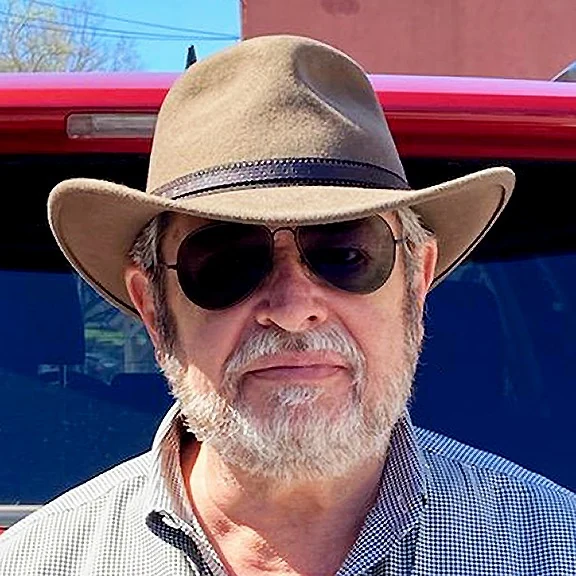 Bob Slitzan, bearded and wearing a wide-brim hat and sunglasses, in front of a red vehicle