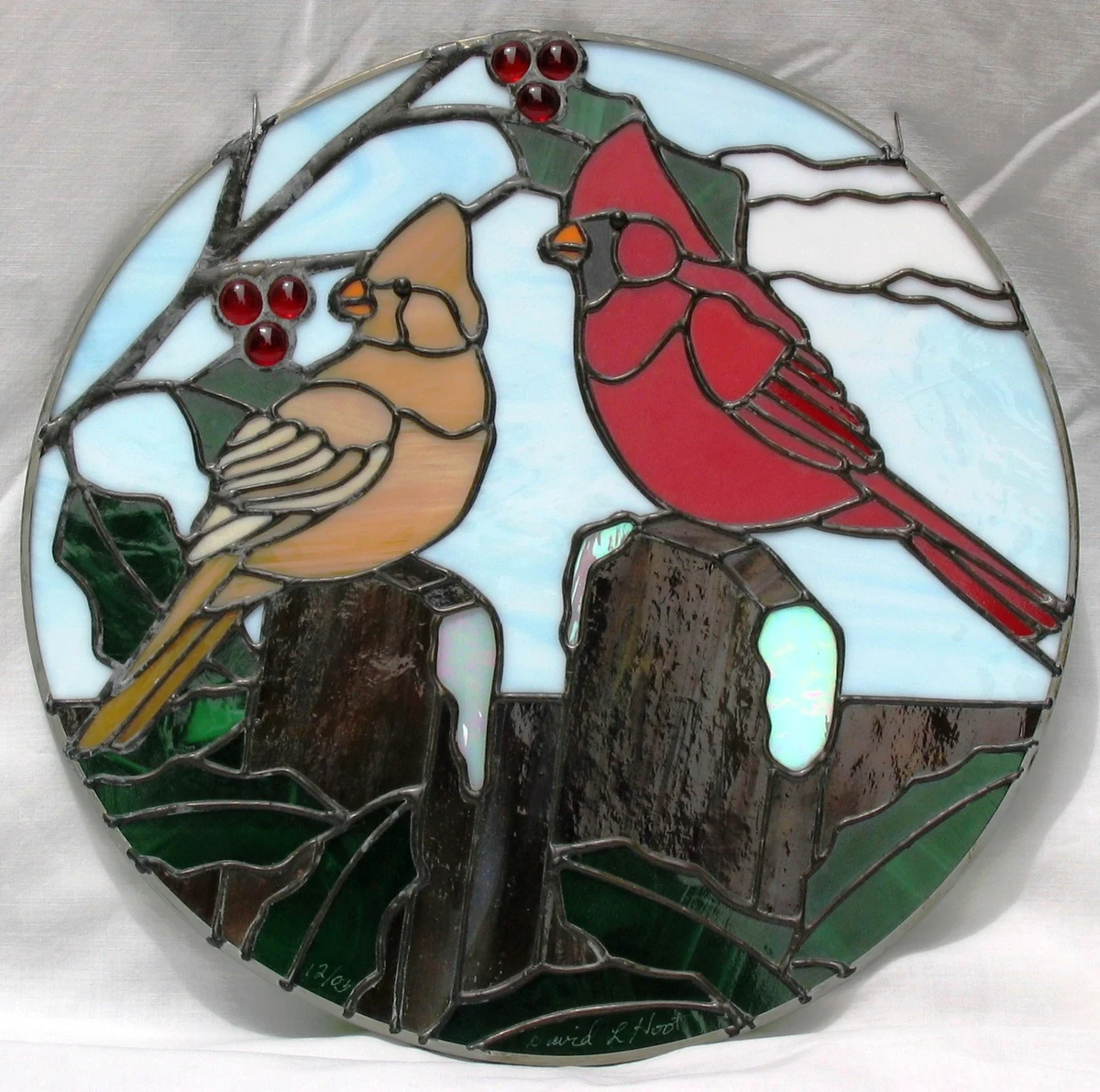 Round stained glass panel of a male and female cardinal perched on weathered fence posts among holly berries and green foliage, against a pale blue sky