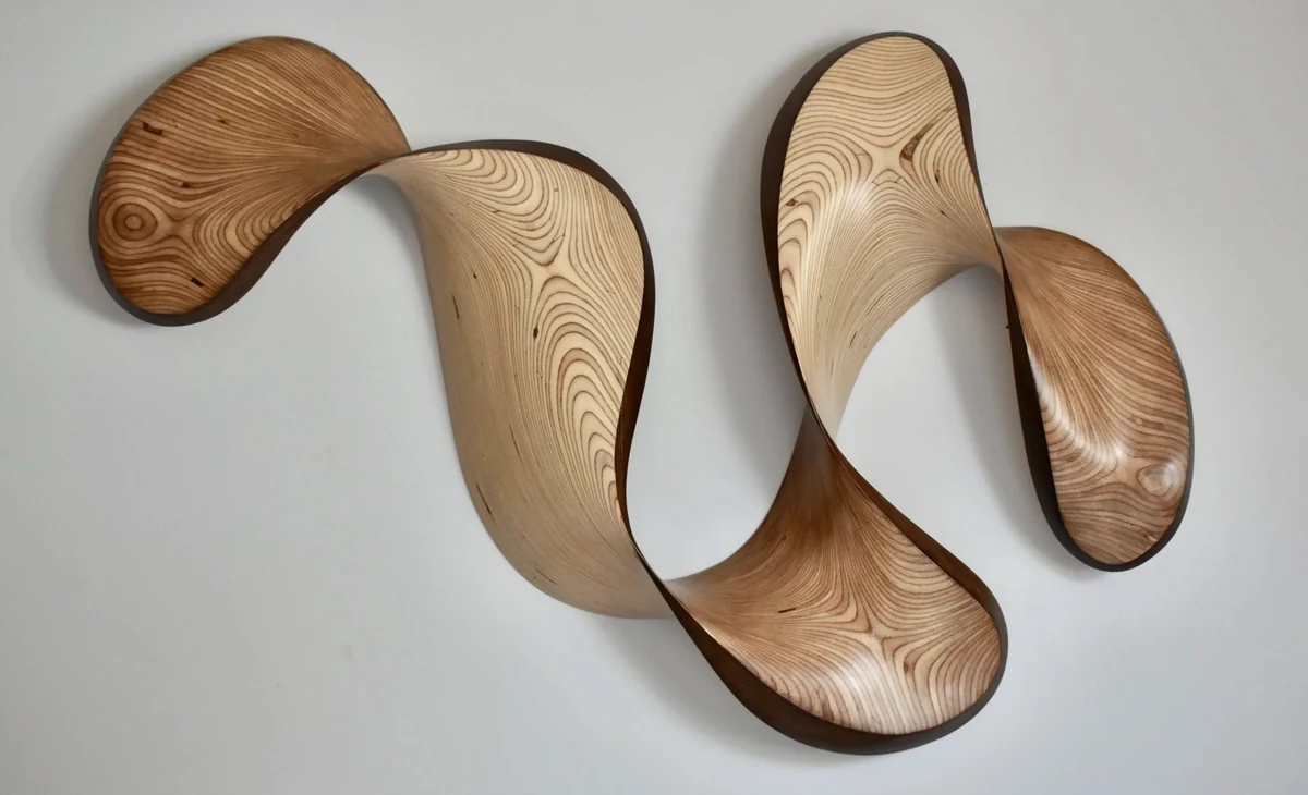 Abstract wall sculpture in natural Baltic birch plywood, a ribbon of undulating curves whose stacked laminations read as concentric contour lines