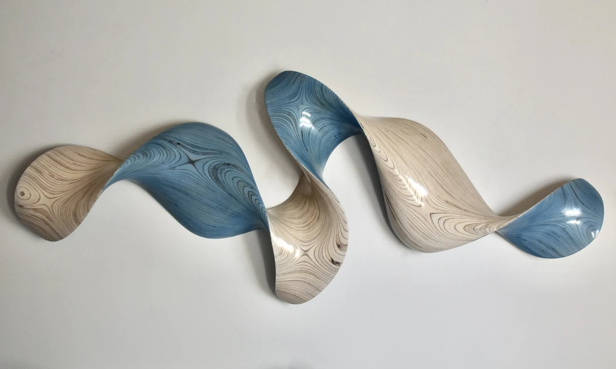 Long horizontal wall sculpture in blue and pale natural wood, a wave form twisting through three crests against a white wall
