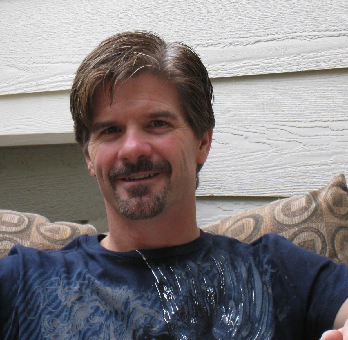 Derrick Crossland, smiling, seated outdoors against white clapboard siding