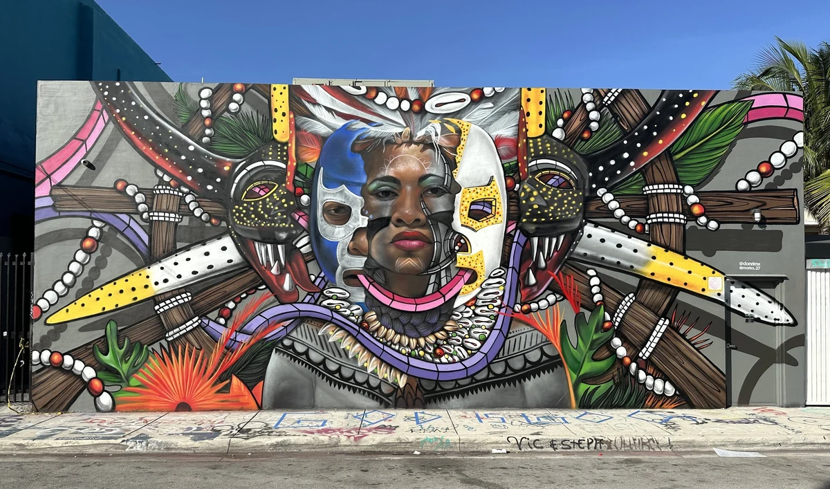 Luchando Por Mi Patria, Wynwood edition: a mural of a woman's face flanked by carved masks, feathers and beadwork on a gray wall