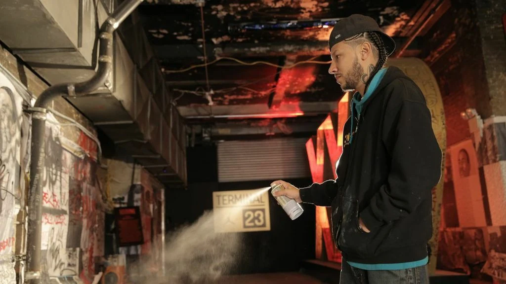 Don Rimx spraying an aerosol can in a warehouse studio, in a black hoodie and cap, painted walls and canvases around him