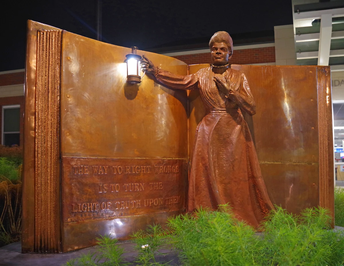 The full Ida B. Wells "Light of Truth" monument at night: a giant open copper book carrying her quotation, with the standing figure raising a lit lantern