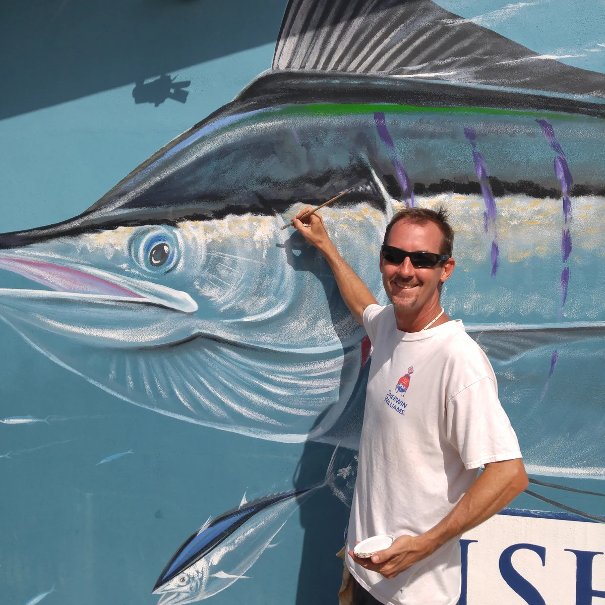 Shannon Paul Wiley painting a large billfish mural, brush in hand and paint cup at his side