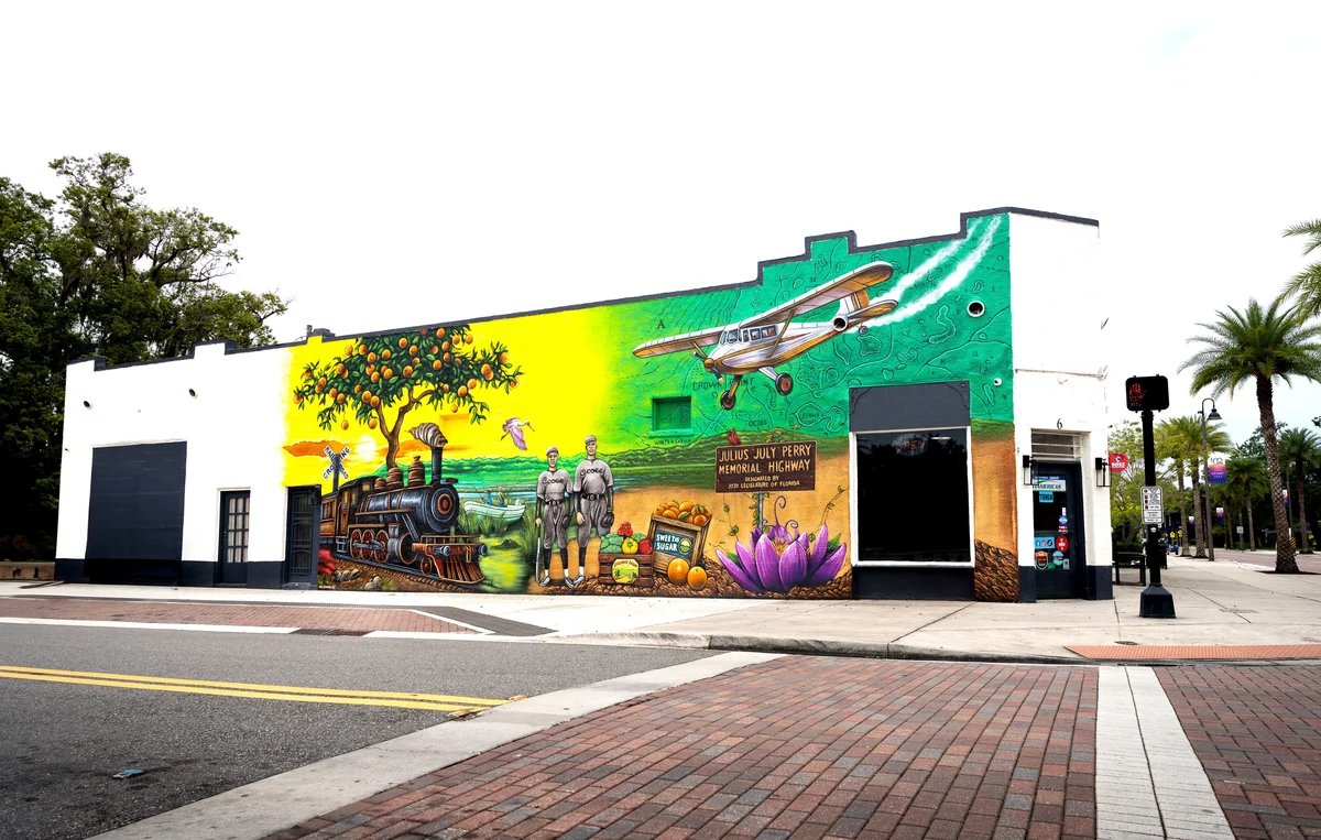 Ocoee Centennial Celebration mural: a steam locomotive, orange grove, biplane and two figures painted across the long side of a downtown building, signed for the Julius July Perry 