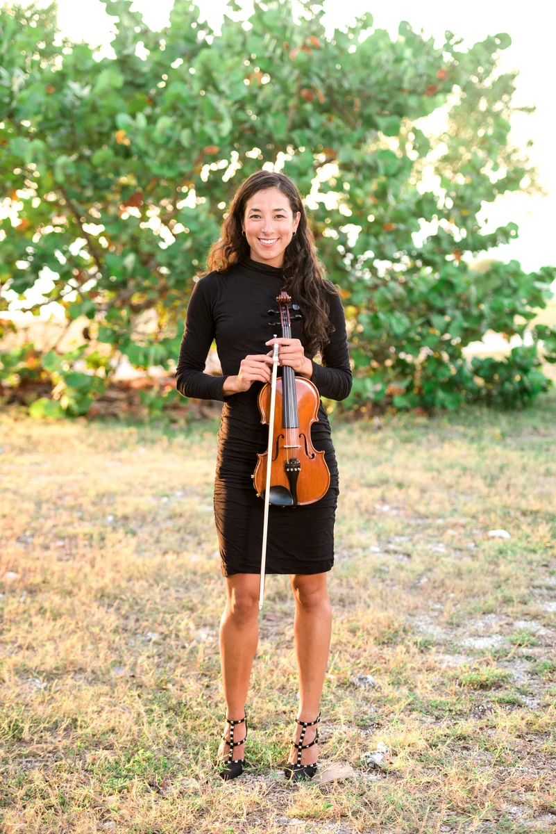 Irie Monte standing full length in a black dress holding her violin and bow, sea grape foliage behind her