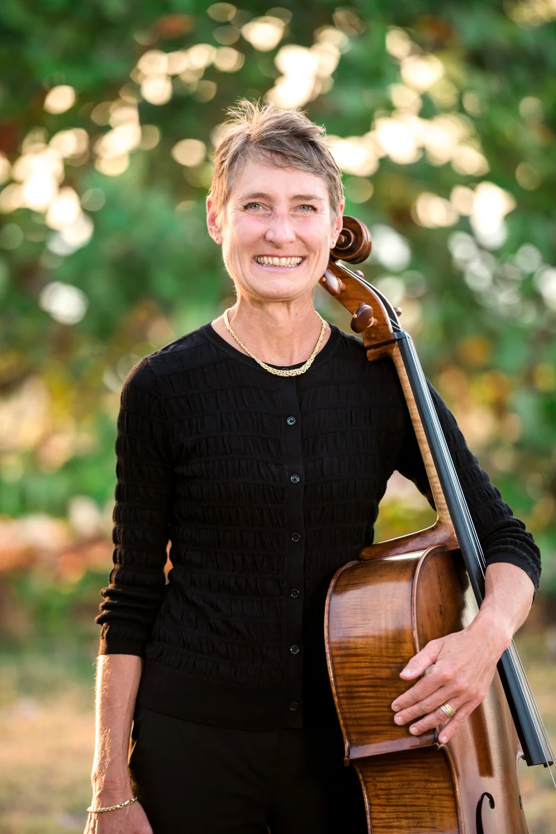 Denise Nathanson in black with her cello resting on her shoulder, green foliage blurred behind her