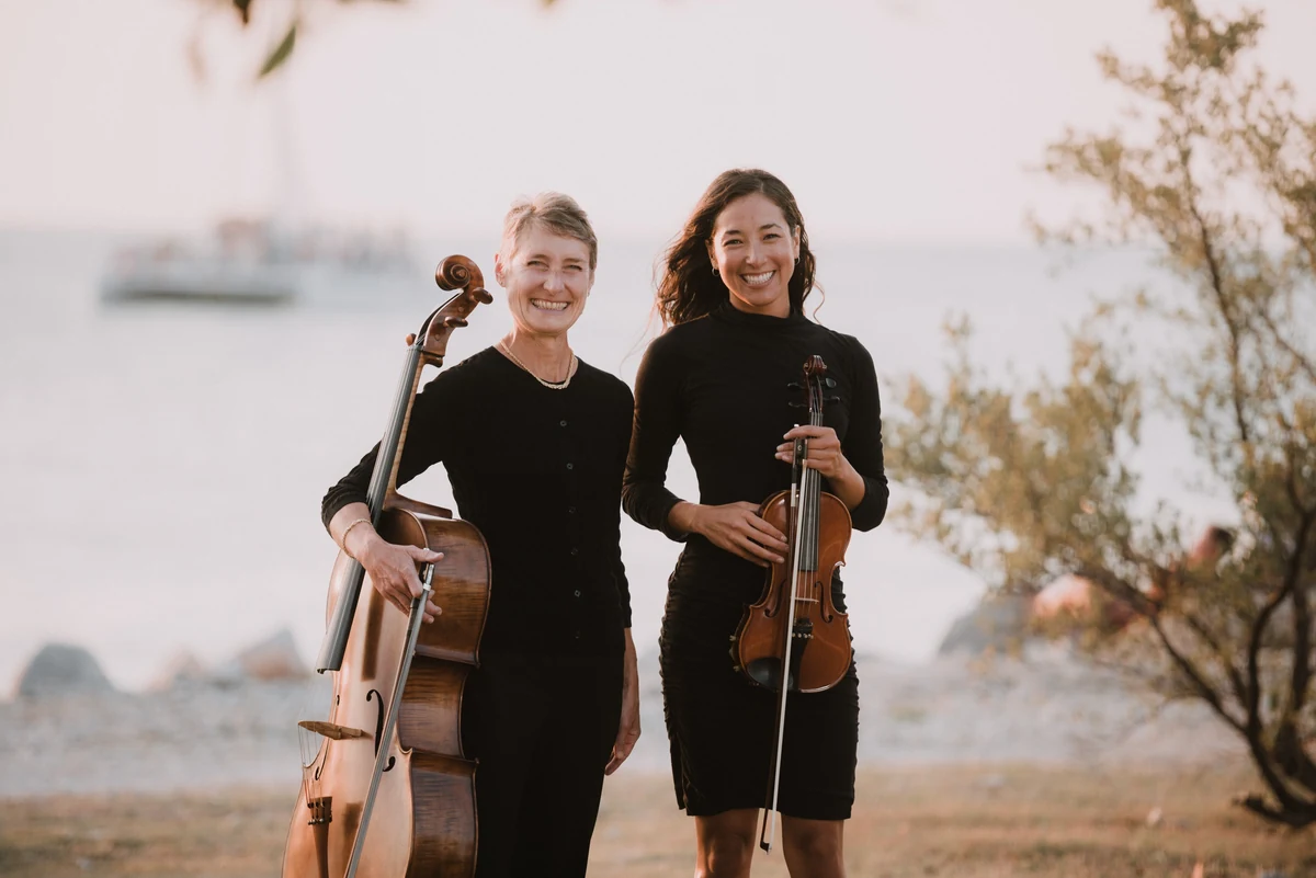 Denise Nathanson holding her cello and Irie Monte holding her violin, standing together in black on a shoreline at dusk