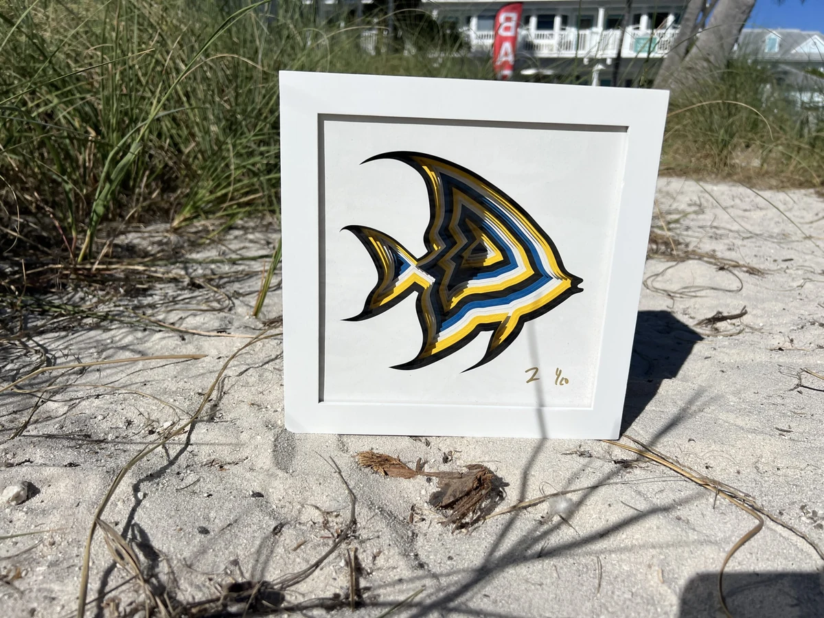 Angel Fish, a layered laser-cut angelfish in black, yellow and blue in a white shadow-box frame, photographed on the sand